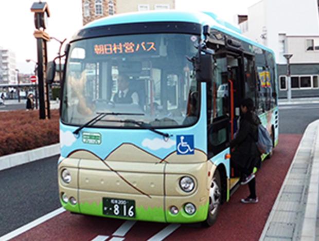 朝日村営バスを右斜め前から撮影した写真。バスの正面には電光掲示板でオレンジ色にて「朝日村営バス」と示されている。バスは全体的に水色のデザインで、正面には黄土色の山と黄緑の草のイラストが描かれている。