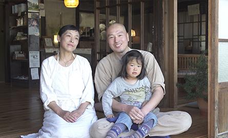 3人の家族を写した写真。木造の家の床の上、上下黄土色の服を着た笑顔の父親はあぐらをかいて娘を抱きかかえており、その左隣で上下白い服を着た母親は正座をして微笑んでいる。