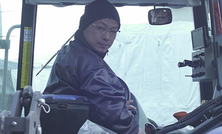 農業用の車の運転席で男性を撮影した写真。男性は紫色のジャンパー、メガネ、紫色のニット帽を身に着け、車を操作している。