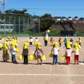グラウンドの上の子供達を撮影した写真。青空の下、同じ黄色の帽子を被り、両手に黄色のボンボンを持った子供達が輪になっている。